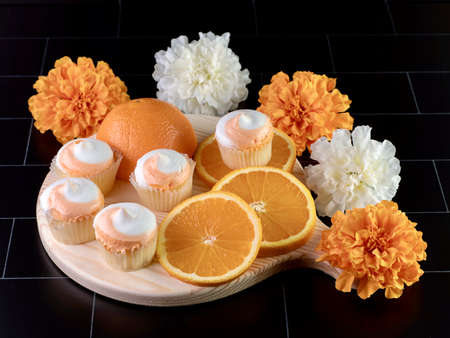 High angle shot of a wooden cutting board on a black subway tile counter with a fresh orange sliced on it and mini cupcakes with orange and white swirled frosting and some artificial flowers.の写真素材