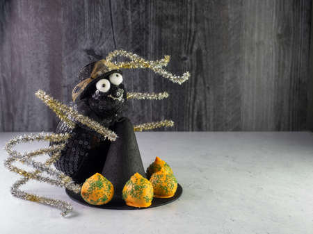 Black spider with big white eyes, a hat and pipe cleaner legs sitting behind a black witch hat with orange and green carrot cake balls lined up on the brim with a black wall behind it.の写真素材