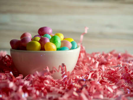 White bowl full of jelly beans, pastel colored and speckled, sitting on pink paper shreds against a...