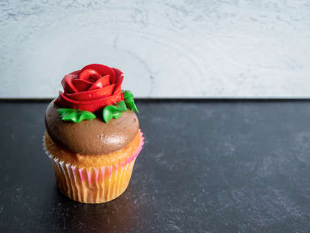 Single vanilla cupcake with chocolate frosting decorated with a red rose and green leaves against a black slate and gray shiplap background, perfect for a birthday, Motherâs Day, or Valentineâs Day!の写真素材