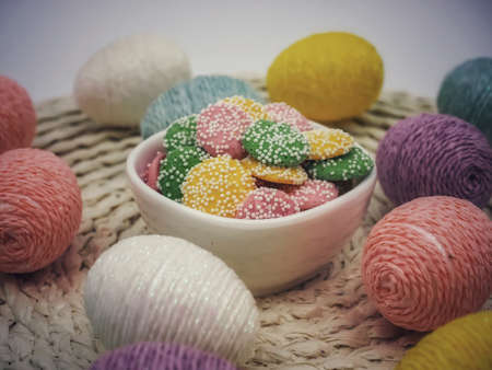 Decorated colorful Easter eggs on a placemat with a small white bowl full of white chocolate colored...