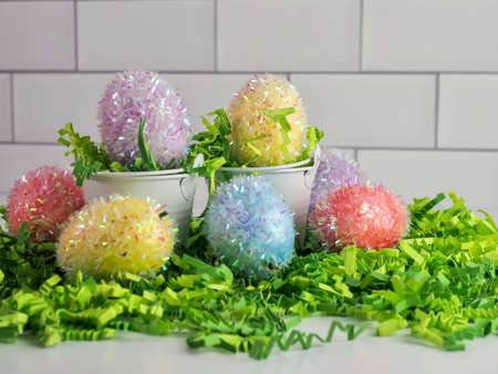 Sparkly pastel colored Easter eggs with two white metal buckets and green paper shreds with a white...