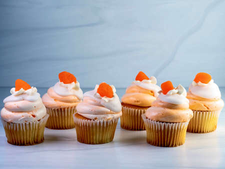Six, 6, yellow cupcakes lined up with orange and white creamsicle frosting swirled up high and an orange gum drop candy on top.  Decadent delicious dessert for a celebration.の写真素材