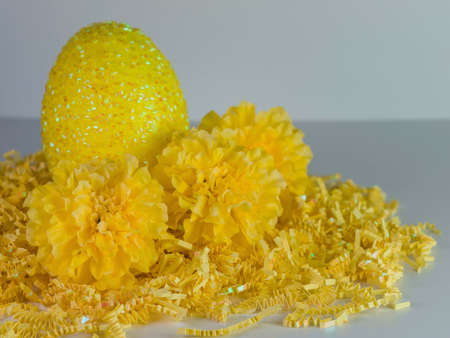 Yellow Easter egg sitting on a bed of yellow paper shreds with yellow carnations on a white...