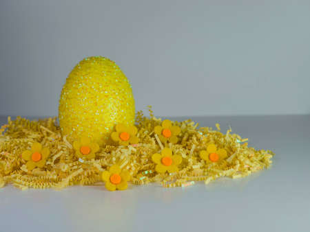 Yellow Easter egg sitting on a bed of yellow paper shreds with little yellow felt flowers on a white...