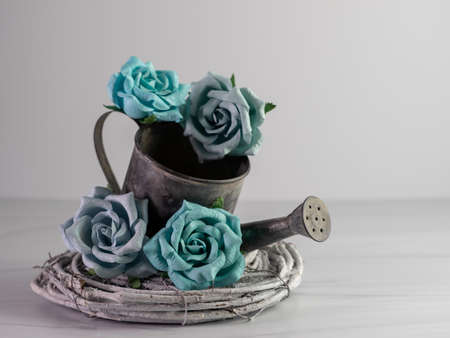 Rustic metal watering can sitting on a gray wooden wreath coiled up with blue and teal paper rose flower blooms on a sandstone background.  Country and simple home decor.の写真素材