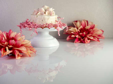 White cake with a frosting flower on top sitting on a white raised cake platter with pink paper shreds decorating it along with two pink dahlia flower blooms all reflecting on a white surface.の写真素材
