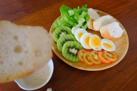 Sliced egg salad serve with vegetable, kiwi, tomato, crispy bread, separated sesame dressing, and bread as foreground (Forty five degree angle view )の写真素材