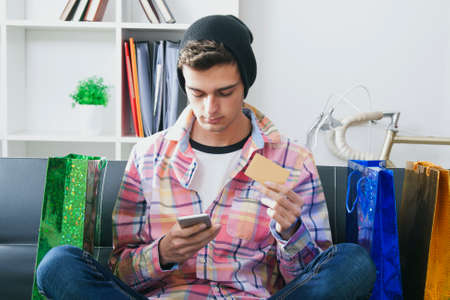 male young with the phone mobile and the card of credit in the sofa of houseの写真素材