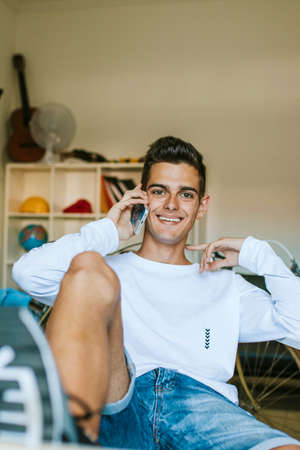 Young man smiling and talking to the mobile phone at homeの写真素材