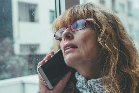 adult or senior woman talking on mobile phone at home in the windowの写真素材
