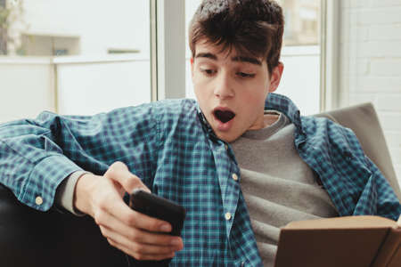 young teenage student with book and mobile phone at home with expression of amazementの写真素材