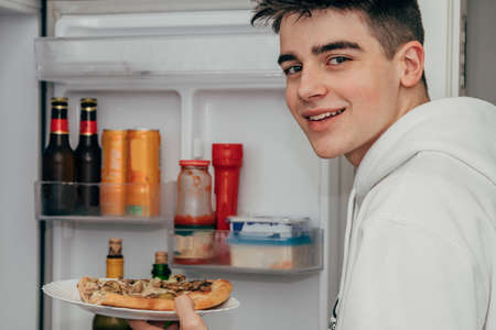 young teenage man at home eating pizza in the kitchen fridgeの写真素材