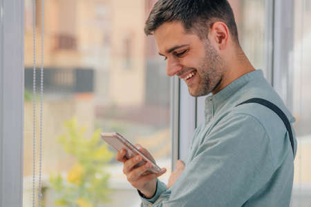 young man looking at the cell phone next to the window smilingの写真素材