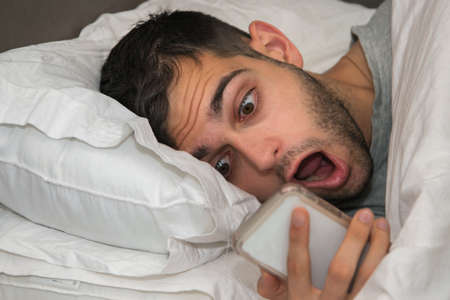 young man in bed staring at the phone, scared or surprisedの写真素材