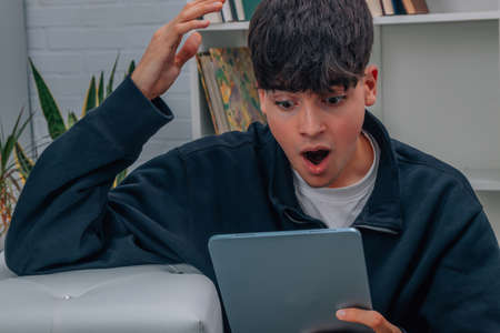 young man at home amazed or surprised looking at computer or tabletの写真素材