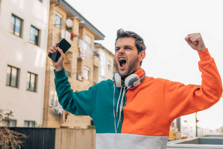 man with phone and headphones in the street excited with joy celebrating success