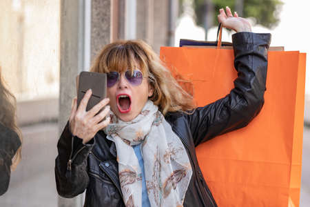 middle-aged woman in the street with shopping bags and mobile phone with a surprised expression