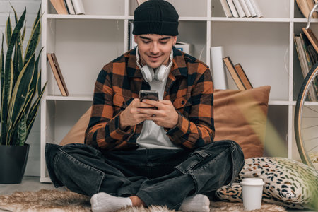 young man at home with mobile phone sitting on the floor wearing hat and autumn winter clothesの写真素材