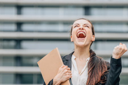 Happy businesswoman with folder celebrating success in front of modern office building