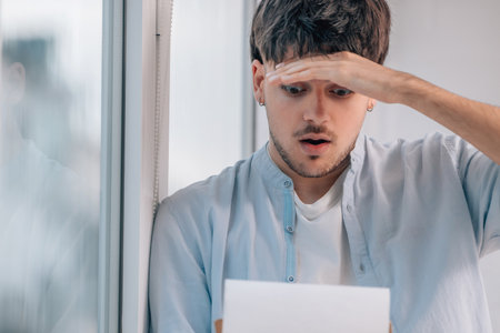 young man with mobile phone indoors next to the window with expression of stress or surpriseの写真素材