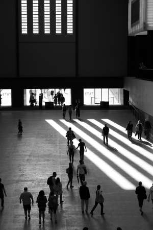 Interior of urban art gallery vast open space with people moving walking, columns of light streaming white as blocks against dark shadow with figures silhouetted in Tate Modern entrance London UKの写真素材