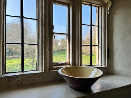 Close up of medieval Elizabethan farmhouse interior window thick hand blown historic glass lwith antique ceramic bowl on sill ledge landscape of grass blue skyの写真素材