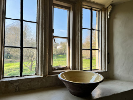 Close up of medieval Elizabethan farmhouse interior window thick hand blown historic glass with antique ceramic bowl on sill ledge landscape of grass blue skyの写真素材