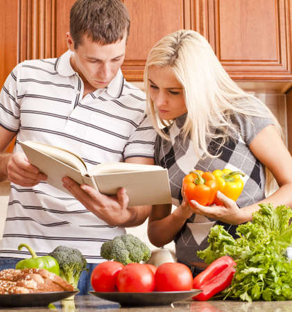 Young couple look at a recipe book in the kitchen. The counter is full of fresh vegetables. Square shot.の写真素材