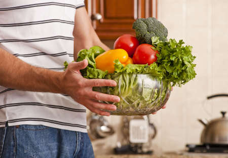 Man in striped shirt holding a bowl of vegetables in kitchen. Horizontal shot.の写真素材