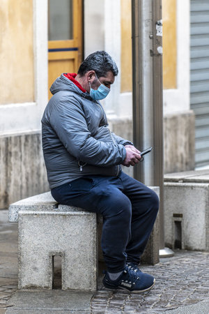 terni, italy jenuary 22 2021: man wearing medical mask sitting on a bench and looking at cellphoneのeditorial素材