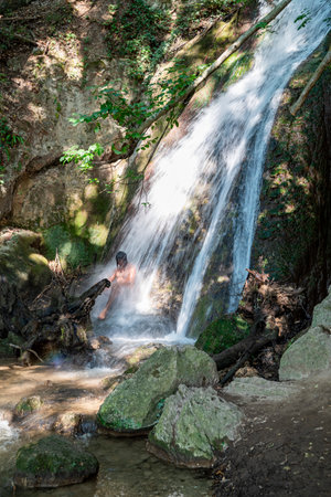 Man under a waterfall in the woods refreshing himself during the summer period.の写真素材