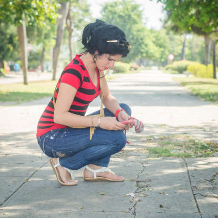 Cute white girl and black hair, look at her cell phone crouched on an old and broken sidewalk, with treesの写真素材
