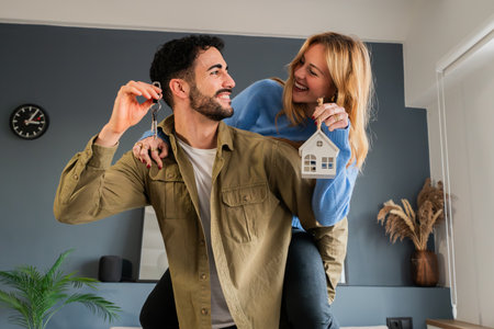 Young married couple enjoying their new house. He is giving a piggyback ride to his wife and they are smiling an laughing holding their apartment keys bonding and with excitement for new beginningsの写真素材