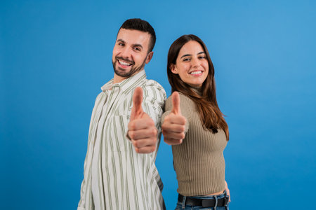 Young adult couple looking at camera with thumbs up in blue background studio. Joyful male and...