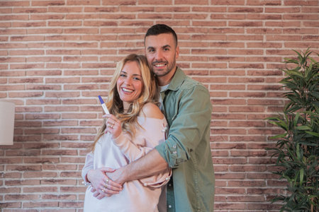 Joyful young wife showing positive pregnancy test result to her husband embracing together while...