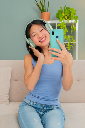 Young chinese girl sitting on the sofa playing music with the smartphone and listening with the...