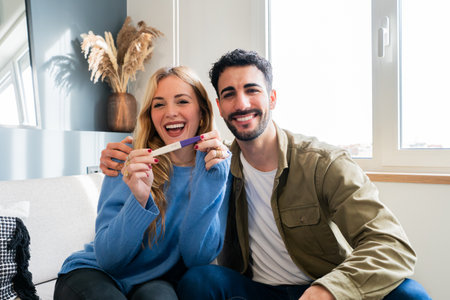 Excited young couple celebrating positive pregnancy test result, smiling and joyful as they embrace the anticipation of parenthood, sitting together in their cozy home, sharing the special moment of happinessの写真素材