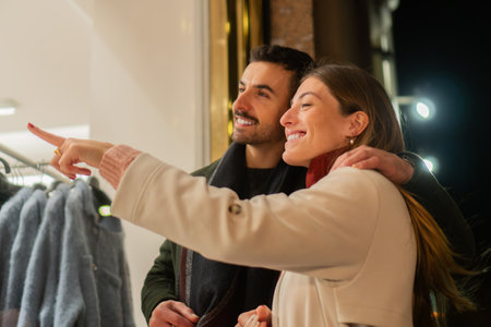 Happy couple window shopping together at night, pointing at store display and enjoying their Christmas holiday shopping experience, sharing a joyful moment in the city during the festive seasonの写真素材