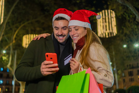 Happy couple shopping online during Christmas time using a smartphone and credit card, enjoying holiday sales, wearing Santa hats, and holding shopping bags in a festive city night sceneの写真素材