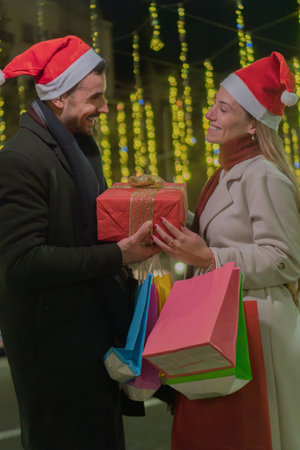 Happy couple wearing Santa hats exchanging Christmas gifts during festive shopping night, surrounded by illuminated decorations, carrying colorful shopping bags, celebrating the holiday season with love and joyの写真素材