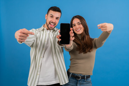 Smiling couple pointing at a mobile phone, showcasing a black display against a blue background, emphasizing technology, communication, and shared enthusiasm for modern devices and connectivityの写真素材