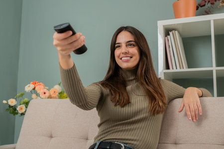 Young woman sitting on a cozy couch, smiling while holding a TV remote in hand, ready to watch television in a modern living room with decorative flowers and shelves in the background.の写真素材