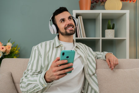 Young adult man enjoying music or a podcast through wireless headphones while holding a smartphone, sitting on a cozy couch in a modern living room, relaxing and smiling peacefullyの写真素材