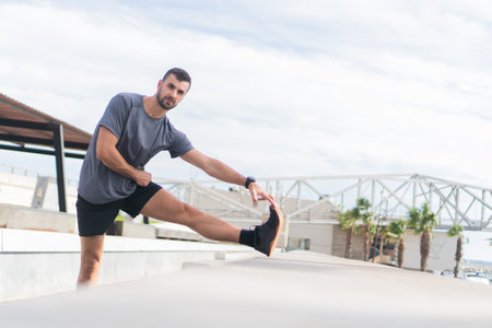 Active Young Man Engaging in Dynamic Stretching Routine to Enhance Flexibility and Improve Overall Fitness While Dressed in Comfortable Sportwear in a Bright Outdoor Area During Daytimeの写真素材