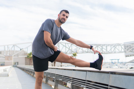 Dedicated Young Man Engaging in Dynamic Stretching Routine to Enhance Flexibility and Prepare for Intense Workout Sessions on a Bright Day by the Waterfront Areaの写真素材