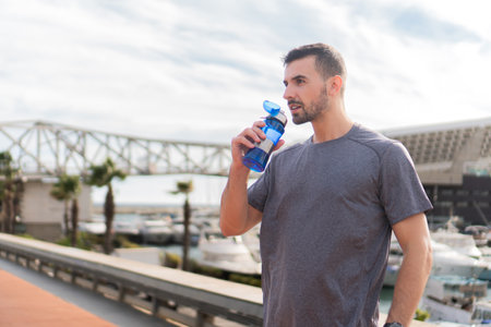 Active Man Quenching Thirst With Refreshing Water During Intense Workout Session In Comfortable Sportwear On A Sunny Day By The Marina Near The Boats And Bridges In A Beautiful Coastal Areaの写真素材