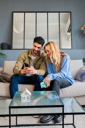 Engaging young couple joyfully browsing smartphones while seated comfortably on a stylish sofa, showcasing their shared interests and connection through technology in a cozy indoor environmentの写真素材