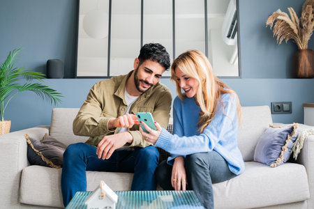 Happy couple of young adults sharing joyful moments while exploring new smartphone features and enjoying each others company in a cozy atmosphere filled with warmth and laughterの写真素材