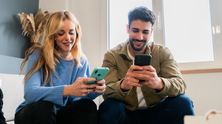 Cheerful young couple engaged in lively conversation while using their smartphones in a cozy indoor setting that fosters connection and enjoyment between them, showcasing modern technology and communicationの写真素材
